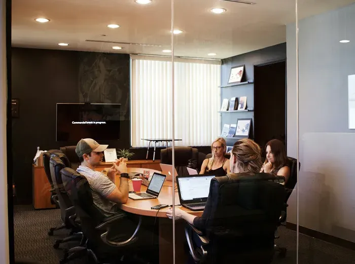 Colleagues around a conference table behind glass walls, discussing plans with laptops, notebooks and coffee cups